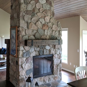May include: A stone fireplace featuring a walnut finish wooden mantel with black metal straps and buttons. The structure is built from multi-colored fieldstones and includes a black glass fireplace door. Text reads: MANTEL FINISH: WALNUT ACCENTS: METAL STRAPS/BUTTONS.