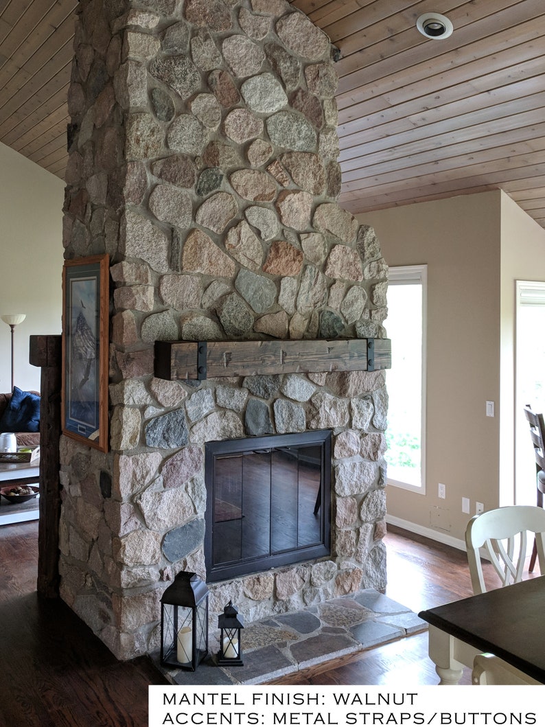 May include: A stone fireplace featuring a walnut finish wooden mantel with black metal straps and buttons. The structure is built from multi-colored fieldstones and includes a black glass fireplace door. Text reads: MANTEL FINISH: WALNUT ACCENTS: METAL STRAPS/BUTTONS.