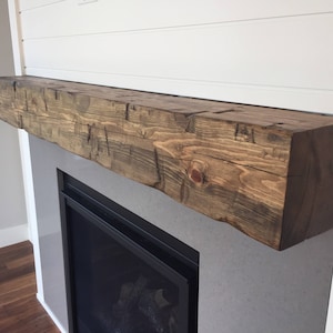 May include: A rectangular fireplace mantel made of dark brown stained wood with a rough, textured surface showing natural grain patterns, knots, and visible cracks. The beam is mounted horizontally above a fireplace insert on a white wall.