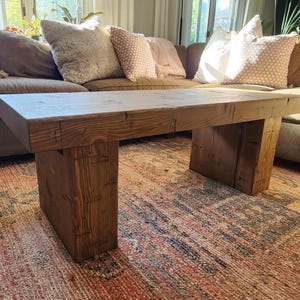May include: A rustic, rectangular coffee table made of dark-stained wood. The table has a thick top and sturdy, block-style legs. The table is approximately 48 inches long, 20 inches wide, and 16 inches high. The table sits on a patterned rug.