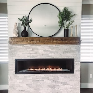 May include: A linear fireplace with a black frame and glowing orange flames, set into a light gray stacked stone wall. Above the fireplace is a thick, dark brown wooden mantelpiece holding a round black mirror, two vases, and two candle holders.