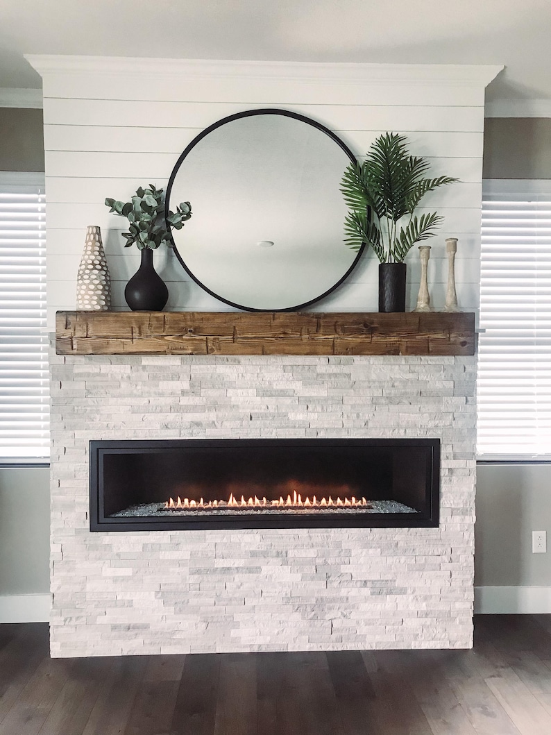 May include: A linear fireplace with a black frame and glowing orange flames, set into a light gray stacked stone wall. Above the fireplace is a thick, dark brown wooden mantelpiece holding a round black mirror, two vases, and two candle holders.