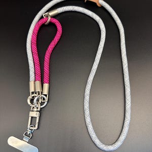May include: A gray and pink lanyard with a silver clasp and phone holder. The lanyard is made of a thick, woven rope material. A small, brown leather piece secures the rope. The pink section is a tightly woven, bright pink rope.
