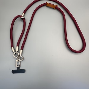 May include: A burgundy phone lanyard with a braided cord design. It features silver-toned metal hardware, a small brown leather accent, and a black phone connector. The lanyard is designed to be worn around the neck.