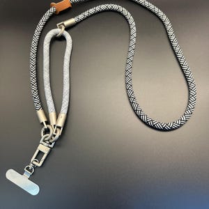 May include: A black and white patterned lanyard with a silver metal clasp and a small brown leather accent. The lanyard is made of a thick, woven rope-like material. The clasp has a rectangular metal plate.