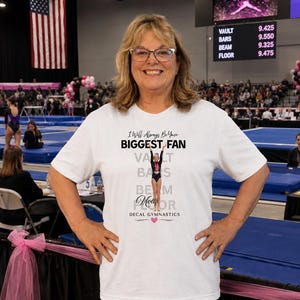 Puede incluir: Una camiseta blanca con el texto "I Will Always Be Your Biggest Fan" y un gráfico de gimnasta. La camiseta también enumera "Vault, Bars, Beam, Floor" con puntuaciones. El fondo incluye un evento de gimnasia y una bandera estadounidense.