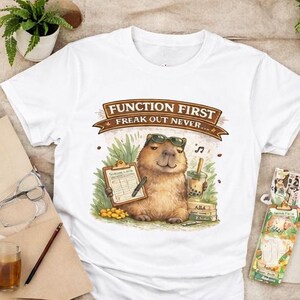 May include: White t-shirt with a cartoon capybara wearing sunglasses, holding a clipboard and bubble tea. The shirt displays the text "FUNCTION FIRST FREAK OUT NEVER" in a banner. The capybara is set against a backdrop of grass and books.