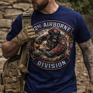 May include: Navy blue t-shirt with a graphic of a gorilla in military gear, the text "2nd Airborne Division" is displayed in a circular emblem. The gorilla is wearing a helmet and combat uniform. The shirt is worn with a cap and camouflage pants.