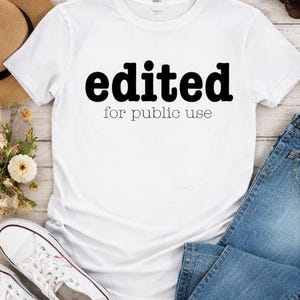 May include: White t-shirt with the text "edited for public use" in black. The shirt is paired with blue jeans, white sneakers, a straw hat, and sunglasses. The items are arranged on a light wood surface.