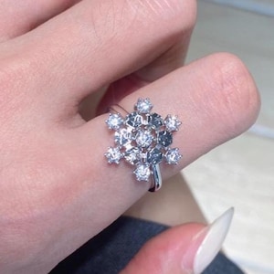 May include: A silver ring featuring a snowflake design. The ring is adorned with multiple clear, sparkling gemstones, creating a brilliant display. The ring is worn on a finger, showcasing its intricate details and elegant style.