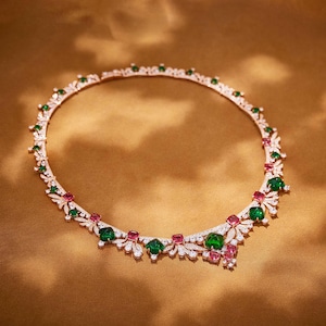 May include: A rose gold necklace with a floral design. The necklace features green emeralds, pink rubies, and clear diamonds. The gemstones are set in a delicate, openwork pattern. The necklace is displayed on a gold background.