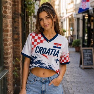 May include: A white V-neck crop top featuring the word CROATIA in bold blue letters. The design includes a red and white checkered pattern, a small Croatian flag, and a blue and white wave graphic. The sleeves and neckline have dark blue trim.