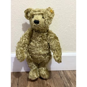 Puede incluir: Un oso de peluche de estilo vintage con un pelaje rizado de color marrón dorado. El oso tiene una nariz negra y una pequeña etiqueta amarilla en la oreja. Está de pie sobre una superficie de madera.
