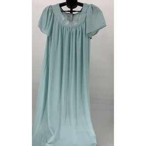 Puede incluir: Un camisón vintage azul claro con mangas cortas y con volantes. El escote está bordado con un delicado motivo floral blanco. El camisón es largo y fluido, hecho de una tela suave y ligera, y cuelga de una percha negra.