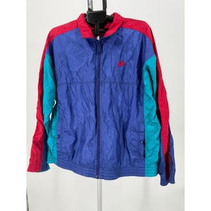 May include: A vintage Nike track jacket in a color block design. The jacket features a blue body with red and teal accents on the sleeves and shoulders. It has a full zipper, a small red Nike logo, and two snap-closure pockets.