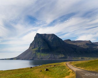 Könnte beinhalten: Landschaftsfotografie mit einem großen, dunklen Berg, der sich aus einem Gewässer erhebt. Eine unbefestigte Straße schlängelt sich entlang der Küste, mit grünem Gras im Vordergrund und einem bewölkten blauen Himmel darüber. Es ist ein Kunstdruck.