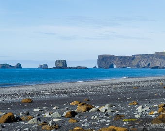 Könnte beinhalten: Eine malerische Küstenlandschaft mit einem schwarzen Sandstrand, blauem Meer und dramatischen Felsformationen. Der Himmel ist klar blau, was eine friedliche Atmosphäre schafft. Das Bild ist eine Landschaftsfotografie.