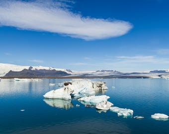 Könnte beinhalten: Eine malerische Landschaft mit einem Gletschersee und schwimmenden Eisbergen unter einem strahlend blauen Himmel. Im Hintergrund sind schneebedeckte Berge zu sehen, und eine große Wolke schwebt über dem Wasser.