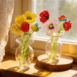 May include: Two small glass vases filled with colorful glass flowers, including yellow, red, and pink blooms. One vase features a small glass bee. The vases sit on a wooden surface near a window with lace curtains.