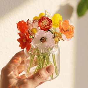 May include: A small glass jar filled with colorful glass flowers in shades of red, orange, yellow, and pink. The flowers have green stems and are held in a clear glass jar. The jar is held in a person's hand.