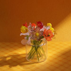May include: A glass jar filled with a colorful bouquet of glass flowers. The flowers are in shades of red, orange, yellow, and white, with green stems. The jar is clear, and the arrangement sits on a yellow surface.