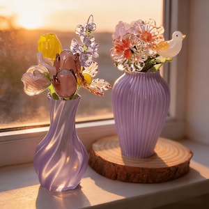 Könnte beinhalten: Zwei Glasblumenarrangements in lavendelfarbenen Vasen. Eine Vase enthält einen Strauß gelber, rosa und lila Blumen. Die andere Vase hat orange und rosa Blumen mit einem Glasvogel. Die Vasen stehen auf einer Holzoberfläche.
