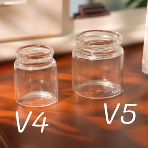 May include: Two small, clear glass jars, labeled V4 and V5, sit side-by-side. The jars have a cylindrical shape with a slightly wider base and a rounded rim. The jars are empty and appear to be made of transparent glass.