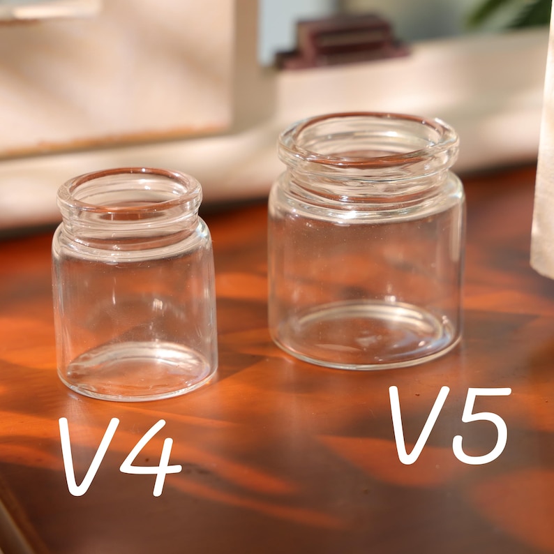 May include: Two small, clear glass jars, labeled V4 and V5, sit side-by-side. The jars have a cylindrical shape with a slightly wider base and a rounded rim. The jars are empty and appear to be made of transparent glass.