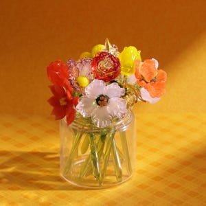 May include: A glass jar filled with a colorful bouquet of glass flowers. The flowers are in shades of red, orange, yellow, and white. The stems are green, and the jar is clear. The background is a solid yellow.