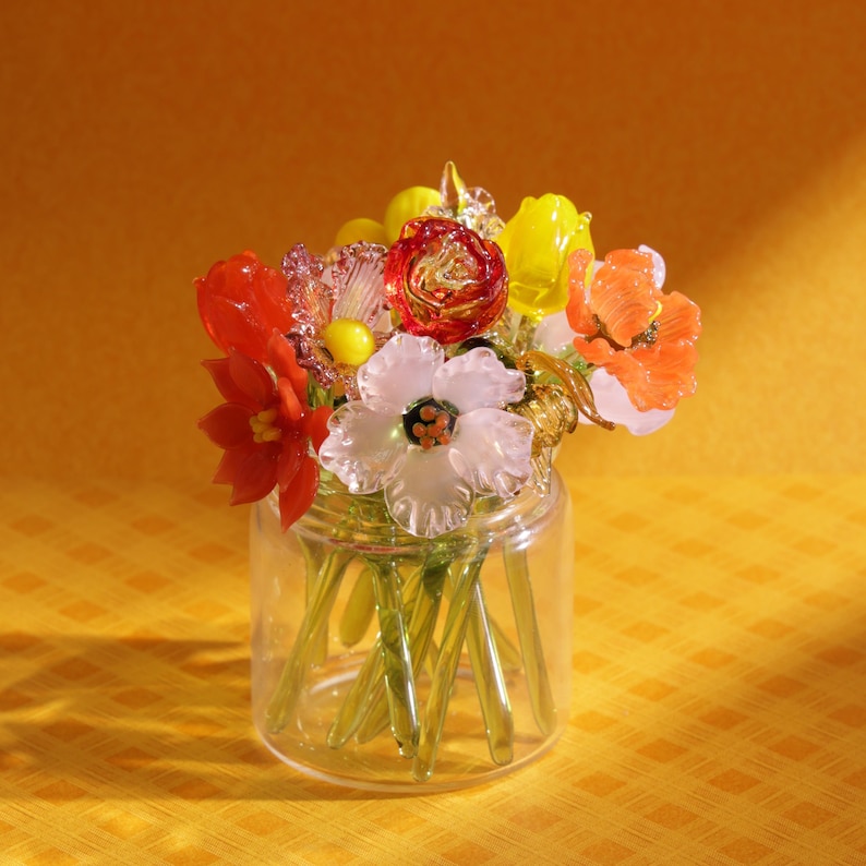May include: A glass jar filled with a colorful bouquet of glass flowers. The flowers are in shades of red, orange, yellow, and white. The stems are green, and the jar is clear. The background is a solid yellow.