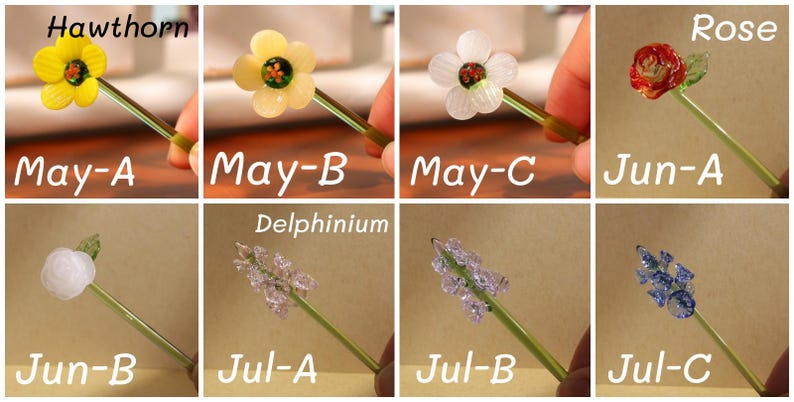 May include: A collection of handcrafted glass flower stems, each featuring a different flower design. The flowers include a yellow Hawthorn, a white flower, a red Rose, and Delphinium. Each stem is held by a hand, with text labels.