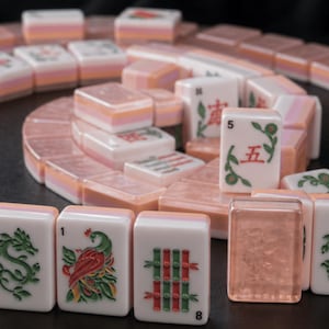 K&ouml;nnte beinhalten: Ein Set rechteckiger Mahjong-Spielsteine, die in einer Spirale angeordnet sind. Die Steine sind wei&szlig; mit durchscheinenden pfirsichfarbenen R&uuml;ckseiten und zeigen eingravierte gr&uuml;ne, rote und schwarze Symbole, darunter chinesische Schriftzeichen, Bambus, Drachen und einen Vogel.