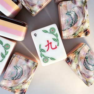 May include: A collection of rectangular mahjong tiles featuring a mix of designs. Some tiles display white faces with red Chinese characters and green floral motifs, while others show iridescent, pearlescent patterns or layered pink, tan, and white bands.