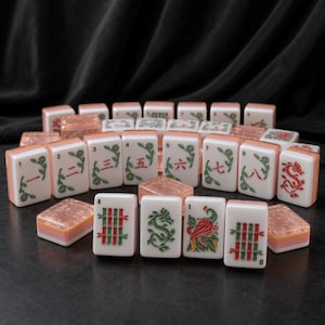May include: A set of rectangular mahjong tiles featuring white faces with green and red engraved designs, including Chinese numerals, bamboo stalks, dragons, and a bird. The tiles have translucent, textured peach-colored backs and sides.