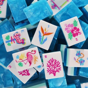 May include: A collection of rectangular mahjong tiles with white faces and translucent blue backs. The tiles feature colorful tropical graphics including a pink flamingo, hibiscus flowers, a bird of paradise, coral, and the word East in script.
