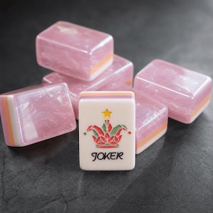 K&ouml;nnte beinhalten: Eine Ansammlung rechteckiger Mahjong-Spielsteine mit einer durchscheinenden, marmorierten rosa Oberfl&auml;che und geschichteten wei&szlig;en und orangefarbenen Unterseiten. Ein Spielstein steht aufrecht im Vordergrund und zeigt ein buntes Narrenkappen-Motiv, einen gelben Stern und das Wort JOKER.