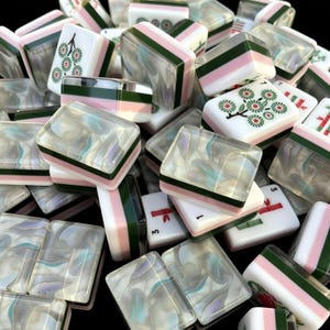 May include: A collection of rectangular mahjong tiles featuring a layered design with a white top, a pink middle band, and a dark green base. Some tiles display green floral patterns or red bamboo symbols, while others have a translucent, marbled finish.