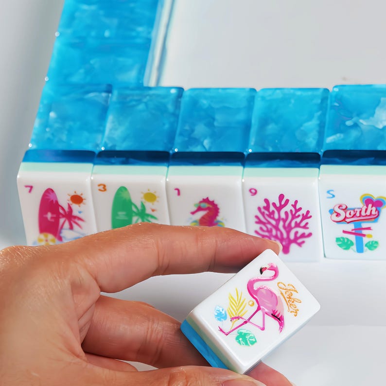 May include: A set of rectangular mahjong tiles featuring a translucent blue marbled top and a white base. The tiles display colorful tropical graphics including a pink flamingo, a surfboard, a seahorse, coral, and the word South in a stylized font.