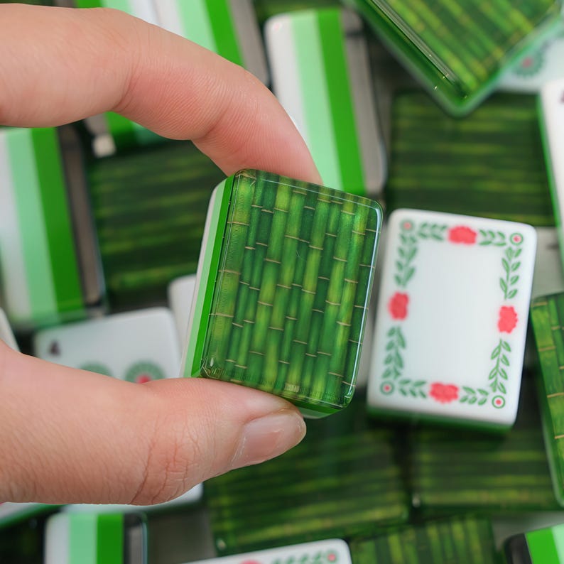 K&ouml;nnte beinhalten: Ein rechteckiger Mahjong-Spielstein mit einem detaillierten Muster aus gr&uuml;nen Bambusstangen auf der Vorderseite. Der Spielstein besteht aus einem glatten, gl&auml;nzenden Material mit wei&szlig;er Basis und Seiten und ist von weiteren Spielsteinen umgeben.