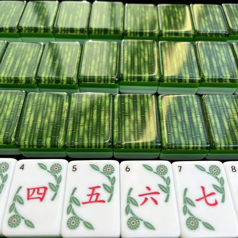 K&ouml;nnte beinhalten: Ein Set rechteckiger Mahjong-Spielsteine mit einem gr&uuml;nen Bambusmuster auf der R&uuml;ckseite und wei&szlig;en Vorderseiten, die die roten chinesischen Ziffern vier, f&uuml;nf, sechs und sieben sowie gr&uuml;ne florale Rankenmotive zeigen.