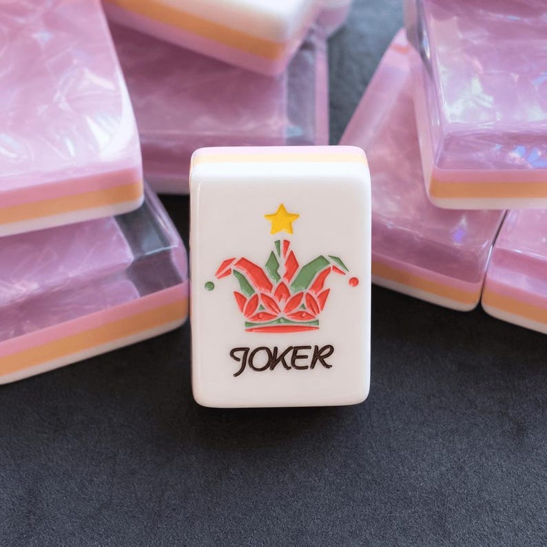 May include: A white rectangular mahjong tile featuring a colorful jester hat graphic with a yellow star above it and the word JOKER printed in black below. The tile has a layered side profile with pink, orange, and white bands.