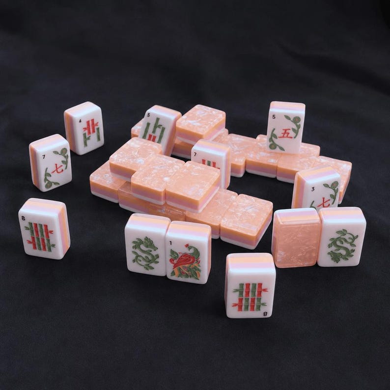 K&ouml;nnte beinhalten: Eine Sammlung rechteckiger Mahjong-Spielsteine mit einer marmorierten pfirsichfarbenen und wei&szlig;en Farbgebung. Die Steine zeigen verschiedene eingravierte Symbole, Zahlen und Illustrationen in roter und gr&uuml;ner Tinte, einige sind unbedruckt.