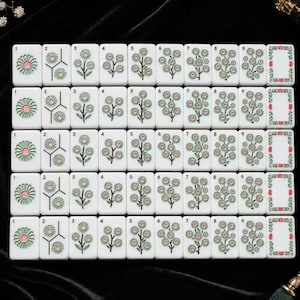 May include: A set of white rectangular mahjong tiles arranged in a grid. Each tile features a green and pink floral design, with numbered tiles from one to nine displaying increasing quantities of flowers, and blank tiles with red floral borders.