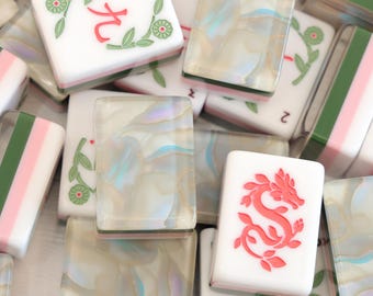May include: A collection of rectangular mahjong tiles featuring a mix of white tiles with red character engravings and green floral accents, alongside translucent, pearlescent tiles with swirling beige, blue, and white marbled patterns.