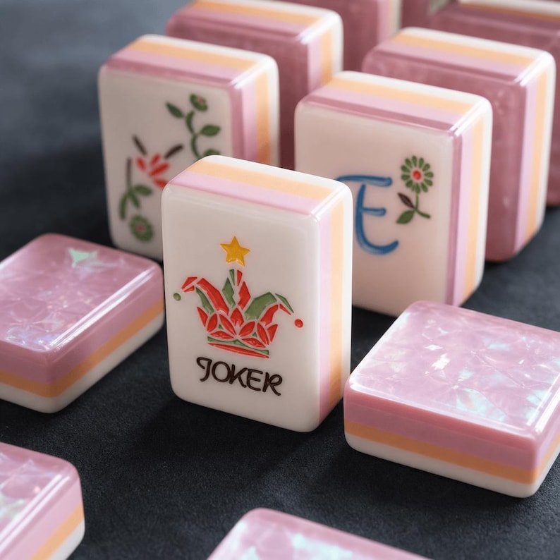 May include: A collection of rectangular mahjong tiles featuring a multi-layered design with white, orange, and pink sides. One tile displays a colorful jester hat graphic with a yellow star and the text JOKER, while others show floral motifs and the letter E.