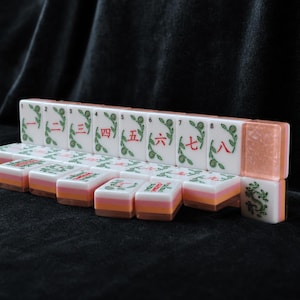 K&ouml;nnte beinhalten: Ein Set rechteckiger Mahjong-Spielsteine mit wei&szlig;en Oberfl&auml;chen, gr&uuml;nen Blumenverzierungen und roten chinesischen Ziffern von eins bis neun. Die Steine haben ein geschichtetes Design mit einer wei&szlig;en Oberseite, einer rosa Mitte und einer orangefarbenen Basis.
