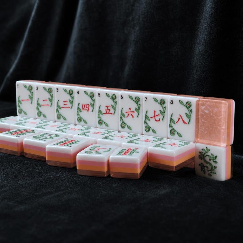 K&ouml;nnte beinhalten: Ein Set rechteckiger Mahjong-Spielsteine mit wei&szlig;en Oberfl&auml;chen, gr&uuml;nen Blumenverzierungen und roten chinesischen Ziffern von eins bis neun. Die Steine haben ein geschichtetes Design mit einer wei&szlig;en Oberseite, einer rosa Mitte und einer orangefarbenen Basis.