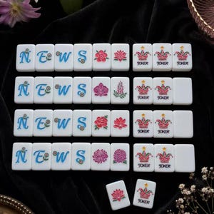 May include: A set of white mahjong tiles arranged in rows. The tiles feature blue letters spelling NEWS, various colorful floral designs in pink and green, and tiles marked with a pink and green jester hat icon and the word JOKER in black text.