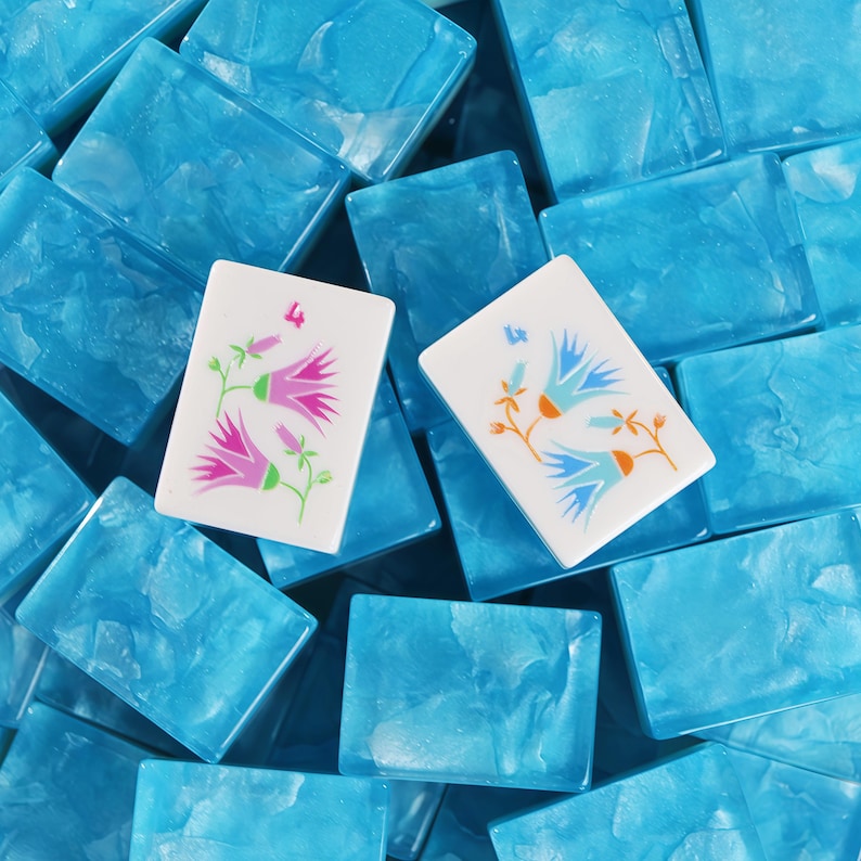 May include: Two white rectangular mahjong tiles featuring floral illustrations and the number 4, resting atop a collection of blue marbled tiles. One tile displays pink flowers, while the other shows blue and orange flowers with green stems.