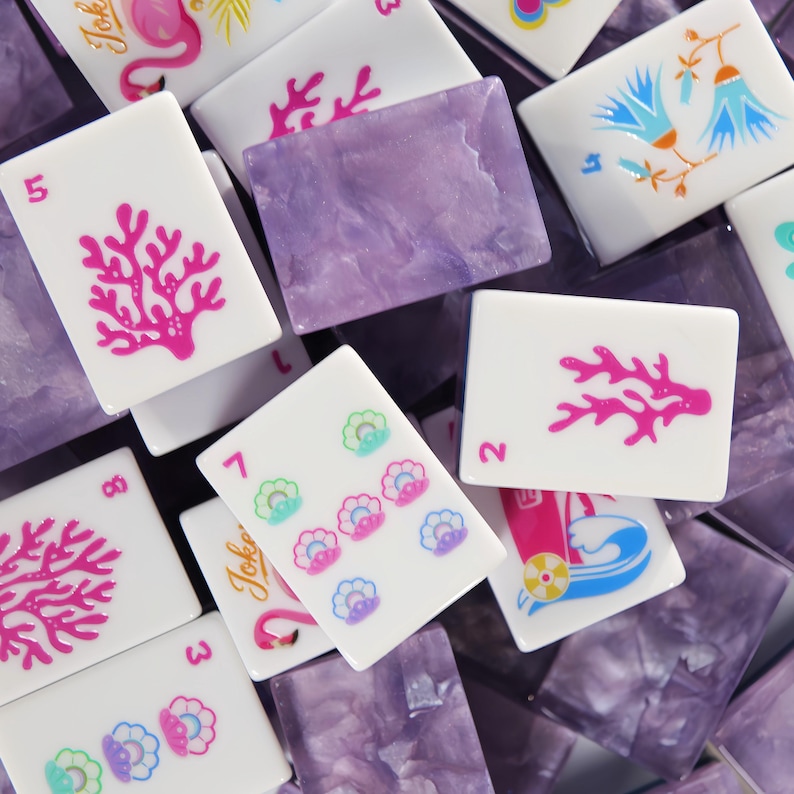 Może przedstawiać: Kolekcja prostokątnych płytek do gry w Mahjong, składająca się z białych awers&oacute;w z kolorowymi nadrukami oraz p&oacute;łprzezroczystych, fioletowych marmurkowych rewers&oacute;w. Białe płytki przedstawiają r&oacute;żne wzory, w tym r&oacute;żowy koral, niebieskie kwiaty, muszle i flamingi.